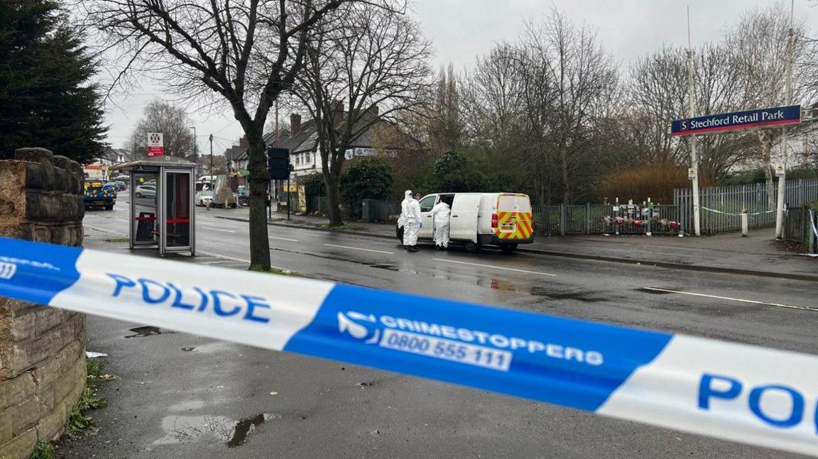 Police tape is in the foreground. A police vehicle is in the background with people dressed in white by it.