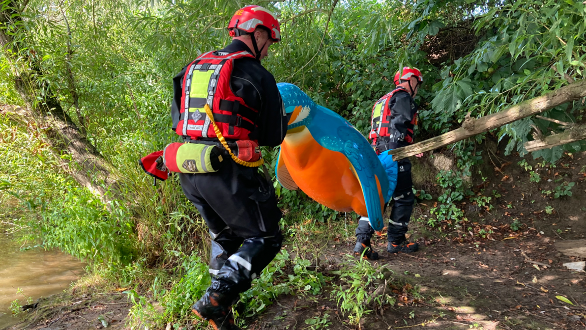 Two firefighters in black and red water rescue gear carrying a large orange and blue penguin sculpture through a wooded area