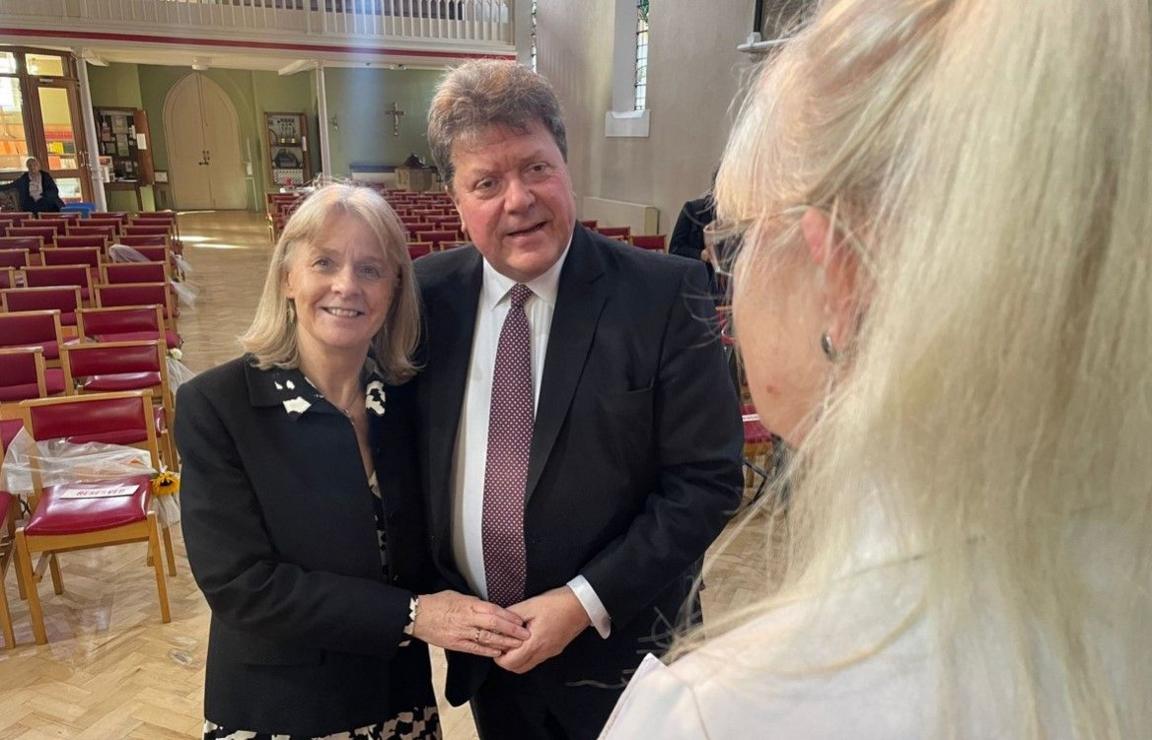 Sue and Alex Tatham are seen over the shoulder of a woman with blonde hair who they are facing. They are holding hands as they stand side by side in a church with empty red seats behind them. There is a light green wall at the back and a white wall to the right.