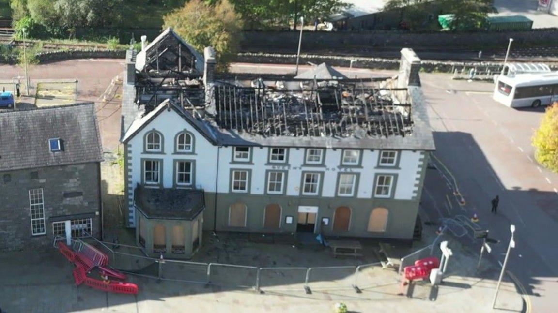 A drone view of the Queens Hotel in Blaenau Ffestiniog a few weeks after the fire in July.  The large detached Victorian building is painted green and white.  But it has been left completely open to the elements with the charred skeletal remains of the roof, devoid of slates and prominent in the photograph.