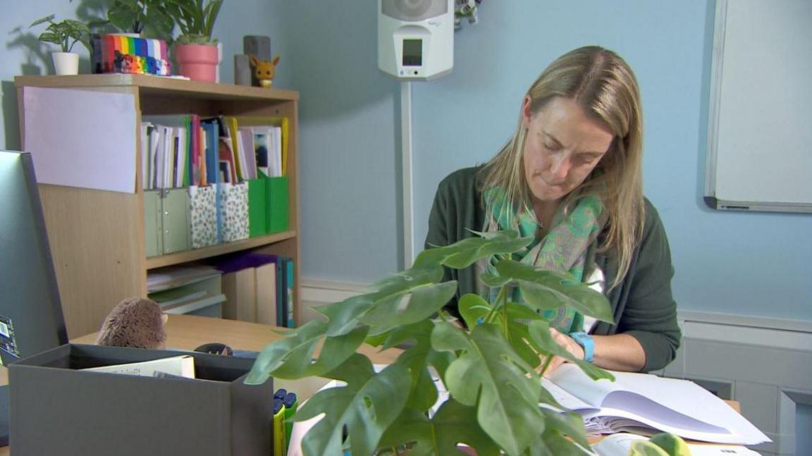 A woman with straight blonde hair sat at a table writing on some paper. She is wearing a green paisley scarf, olive cardigan and white blouse. She is sat at a desk with a green plant in the foreground. Behind her to the left are some shelves with papers in folders. To the right is a white board.