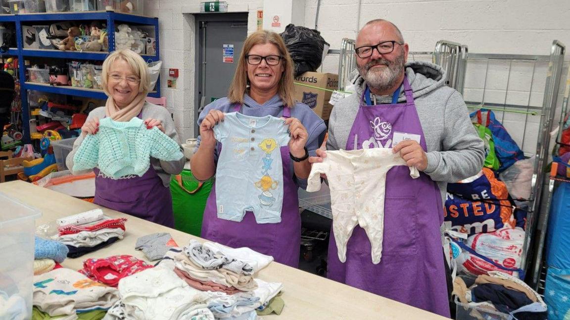 Two women and a man from the charity smile at the camera while holding baby clothes. They are wearing purple and all three have glasses. There are piles of baby clothes on the table in front of them. Behind them, there are shelves with toys. 