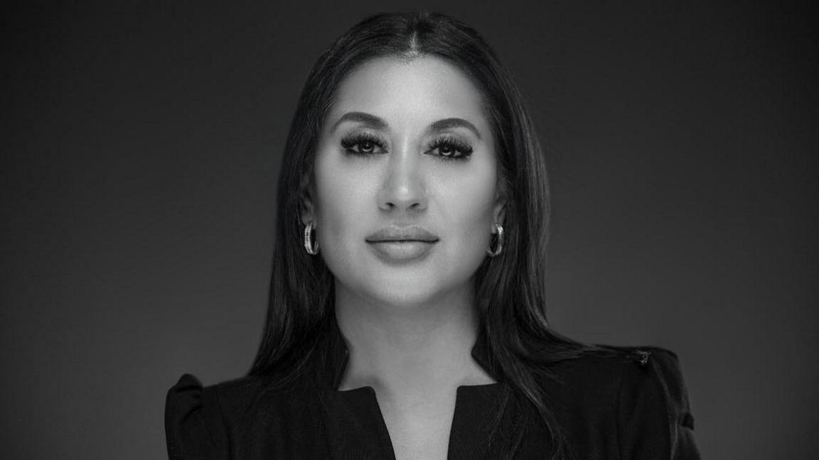 Miriam Altaf with long dark hair and circular earrings wearing a black top looking at the camera against a black background.