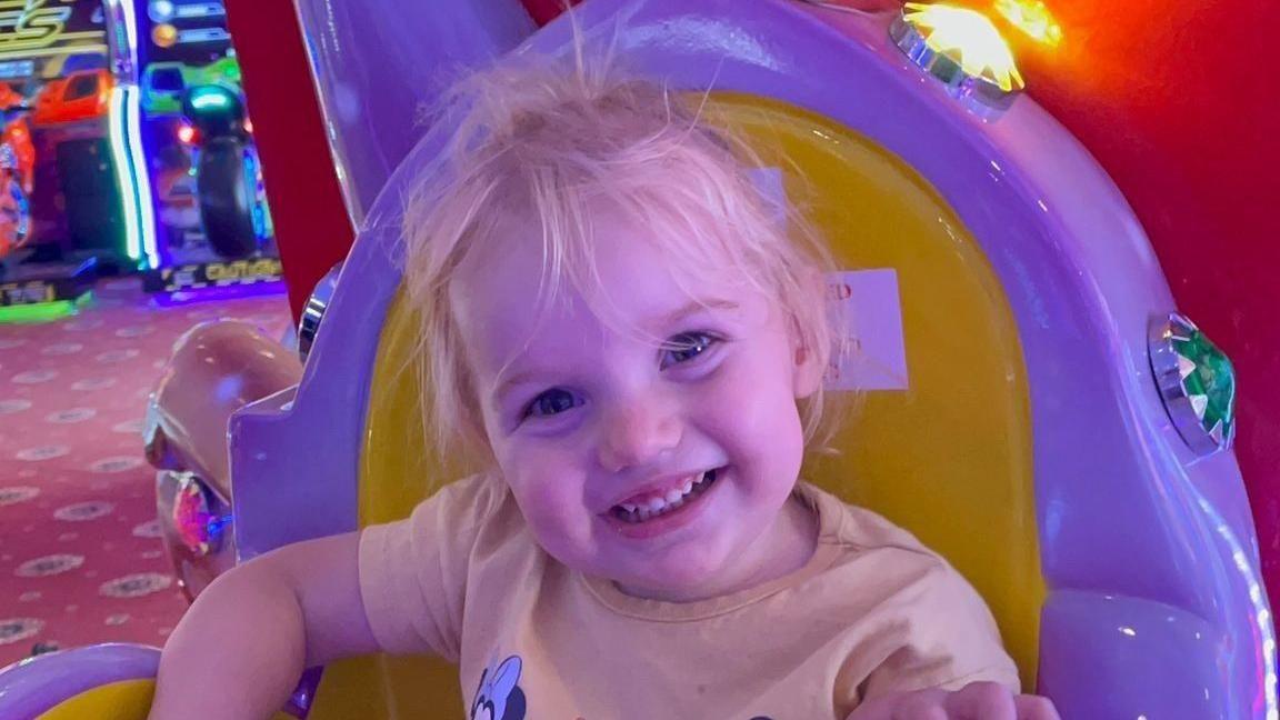 Isabella Jonas-Wheildon, a young blonde toddler wearing a yellow t-shirt smiling at the camera. She is sitting on a yellow and purple chair at an arcade.