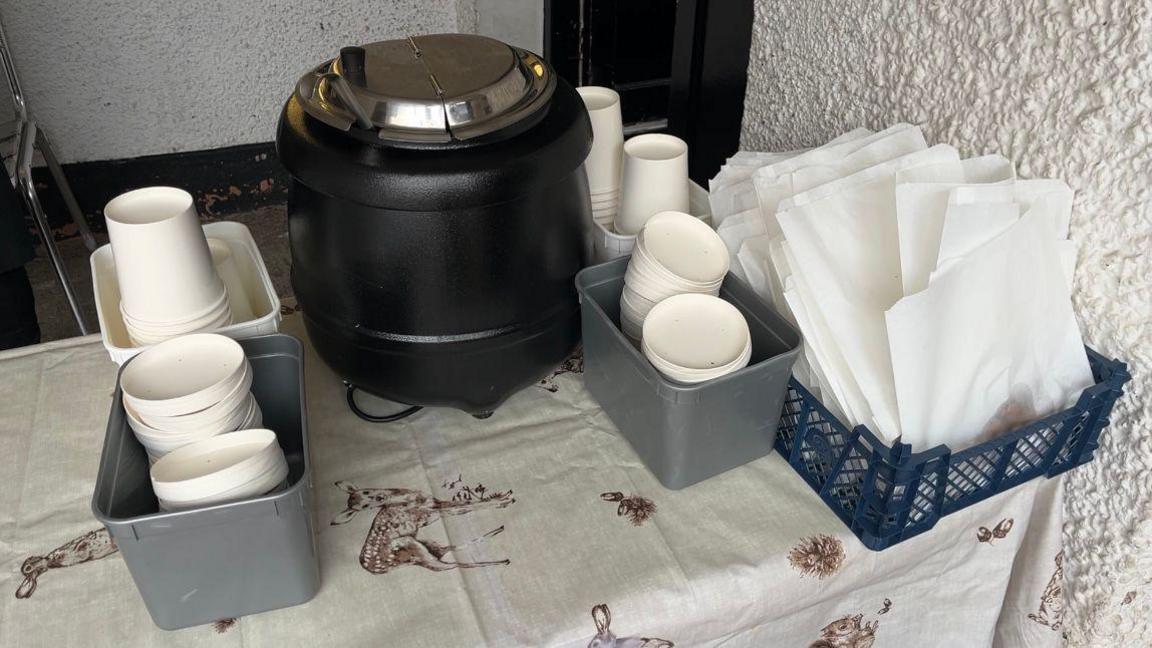 A table set up outside, with paper bags full of croutons on the right of the shot, with a soup cauldron in the middle. Surrounding the cauldron is paper cups and lids.