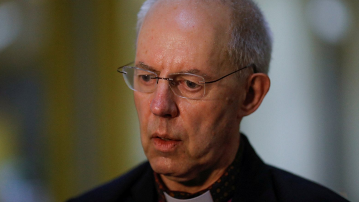 Justin Welby, wearing thin-framed spectacles, a dog collar and a black jacket, bears a serious expression as he speaks during a press interview