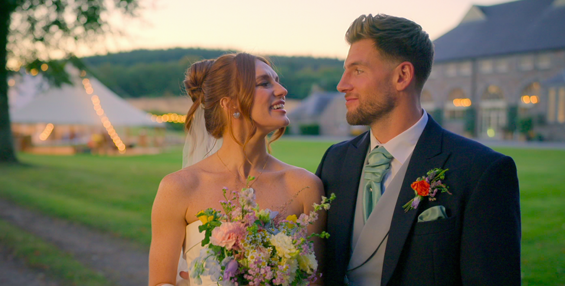 Megan and Kieran looking at each other smiling, wearing a wedding dress and suit respectively. A large countryside house can be seen in the background.