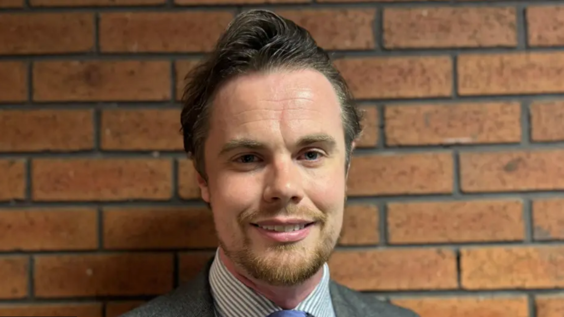 Councillor Philip Faircloth-Mutton is smiling at the camera he is wearing a suit and tie, with a striped shirt. He  is standing with against a brick wall 