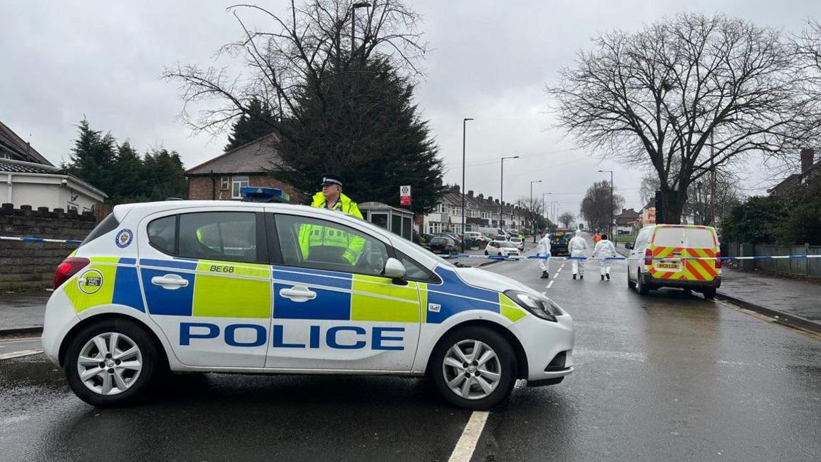 A police car is in the foreground near police tape going across the road. Behind the tape is another police vehicle and three people wearing all white are near it.