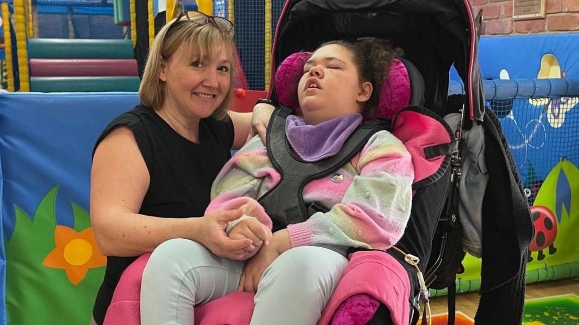 A lady is keeling beside a teenage girl in a supported wheelchair and holding her right hand. The woman has short blonde hair with glasses propped on top of her head and is wearing a black sleeveless top. The girl is in a multi-coloured jumper of pink, pastel blue and pastel green. She has dark curly hair and has her eyes closed. Behind the pair, brightly coloured soft play barriers can be seen, decorated with a ladybird and a butterfly.