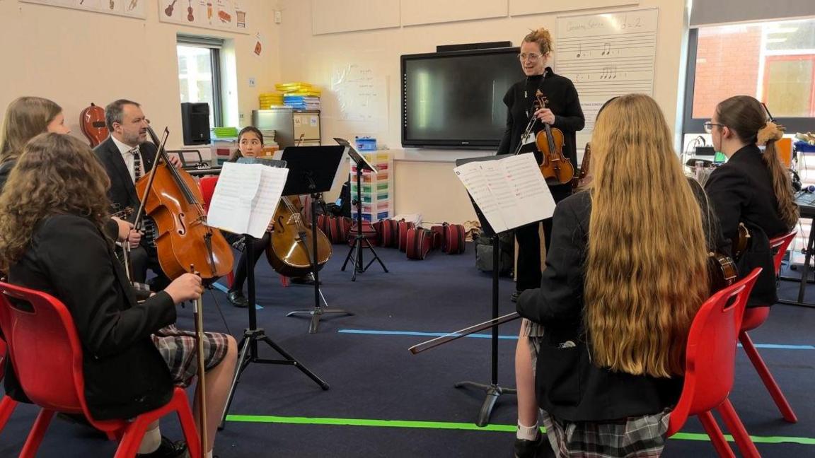 Children playing various string instruments are in a semi circle in a music classroom. They're sat on a red chairs facing away from the camera looking at Mrs Perre, the teacher holding a violing, in the middle.