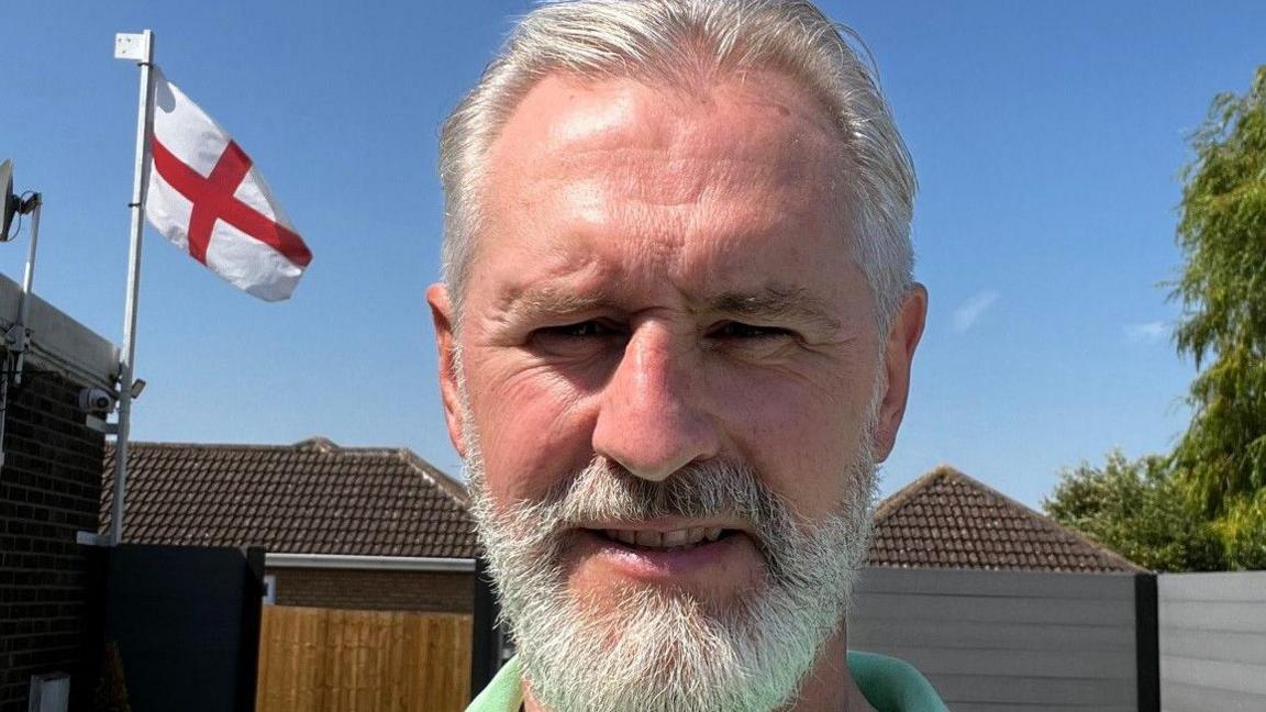 A close up of Martin Skilbeck, wearing a green top and who has a white beard. He is photographed outside and an England flag can be seen in the background. 