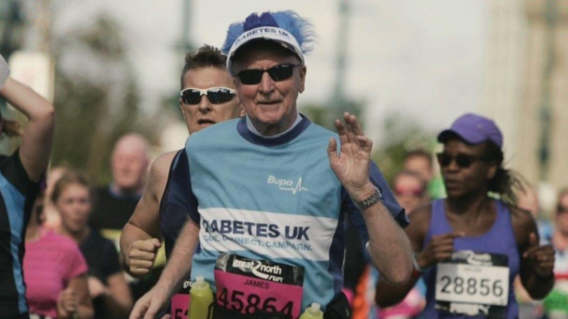 A Jim Meehan wearing dark black glasses and a light blue running jersey with a pink sticker with the numbers 45864. He is also wearing a white hat with fluffy blue hair sticking out of the top.