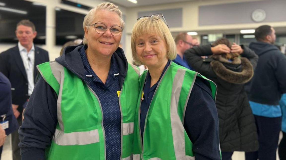 Two women wearing green hi-vis sleeveless jackets. The woman on the left has short hair and glasses. The woman on the right has a blonde bob and has her glasses perched on top of her head.