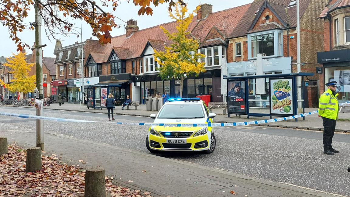 A police officer standing on the taped-off Banbury Road, standing in front of a police car. The car is parked behind blue and white police tape. 