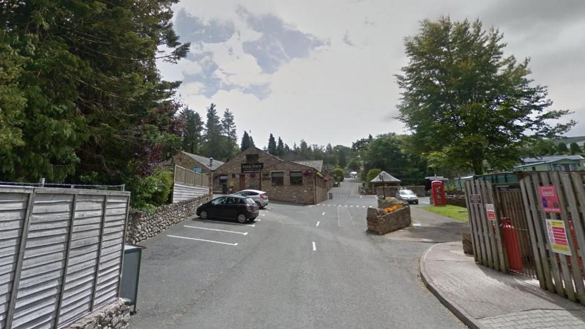 The entrance to Park Foot Holiday Park. A large, single storey stone building sits alongside a long drive. Cars are parked nearby and an old red telephone box is on the right hand side of the drive.