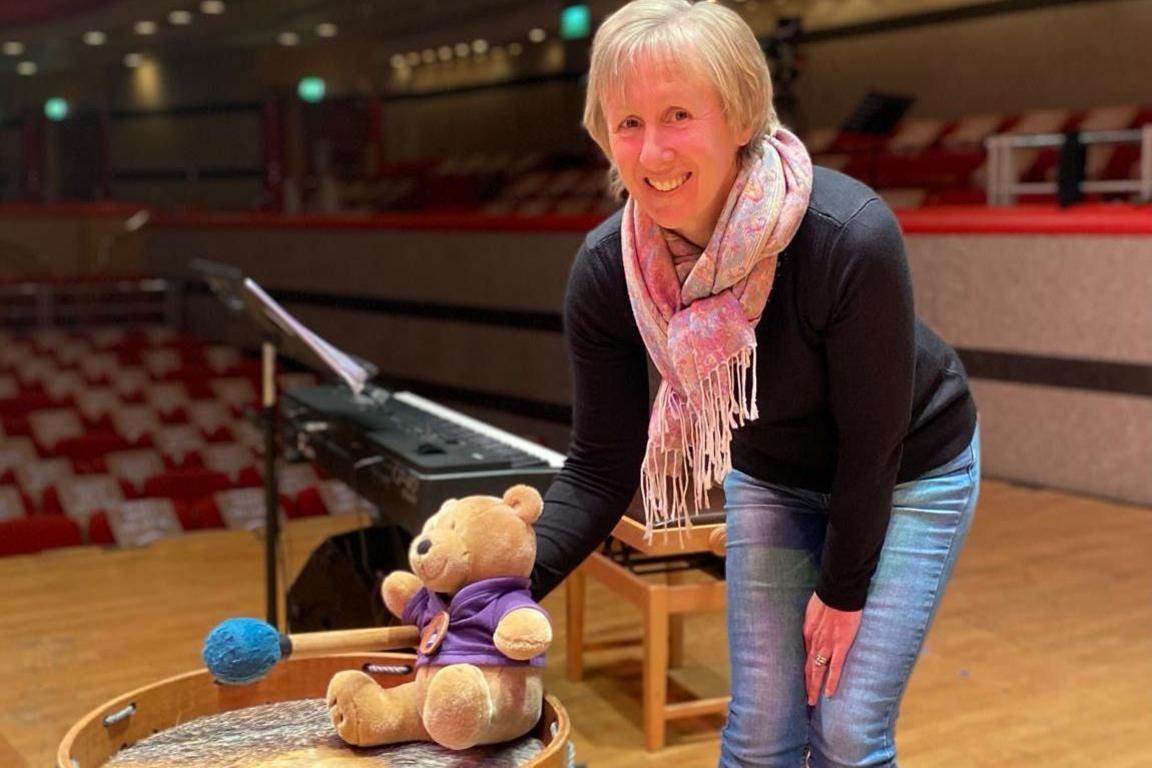 A woman stands in a hunched over position holding a teddy bear that is sat on a drum. She is standing on a stage of an empty hall, behind her is a keyboard and further in the background are rows of empty seats