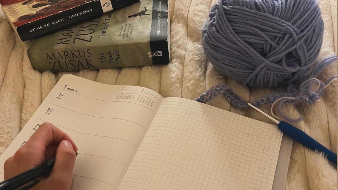 A partial hand, holding a pen, poised over an open page of an empty diary. In the top right is a ball of wool with a crochet needle, and two books in the top left.