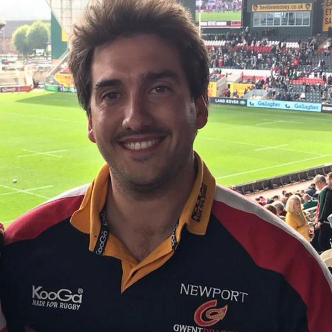 Ryan PRice, who has brown eyes and brown hair, pictured at a rugby match in a Newport Gwent Dragons (the former name of Dragons) jersey, with a yellow collar, red shoulders and black body