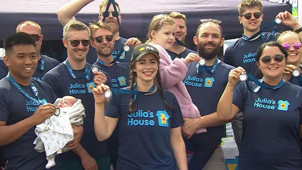 A group of people with blue T-shirts reading Julia's House are smiling at the camera, showing off medals. They appear to be in front of a purple tent