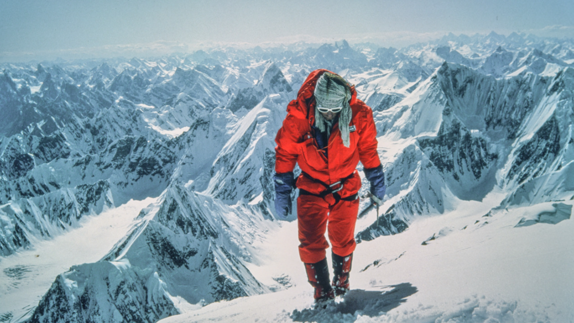 Photograph from the 1975 British Mount Everest Southwest Face expedition. A man in bright-orange hiking gear, sunglasses and gloves walks up towards the camera. Snow is all around him 