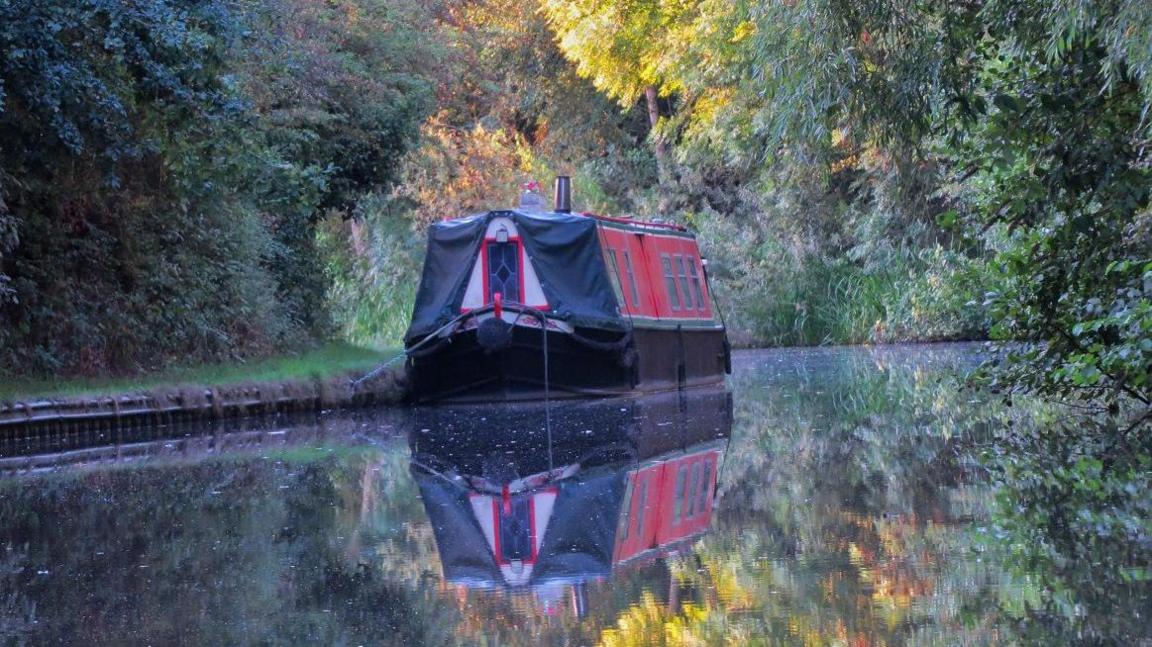 A canal boat sits on the water with trees overhead, some changing colour for autumn. They are reflected in the water.