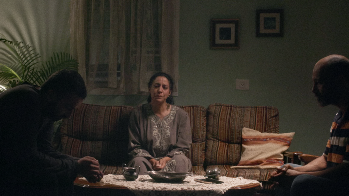 A scene from Iris Prize 2024 winning film Blood Like Water, with an upset-looking woman sitting on a sofa in a room, with two men in shadows sitting on either side of her at right angles, one of them looking down, and the other looking ahead