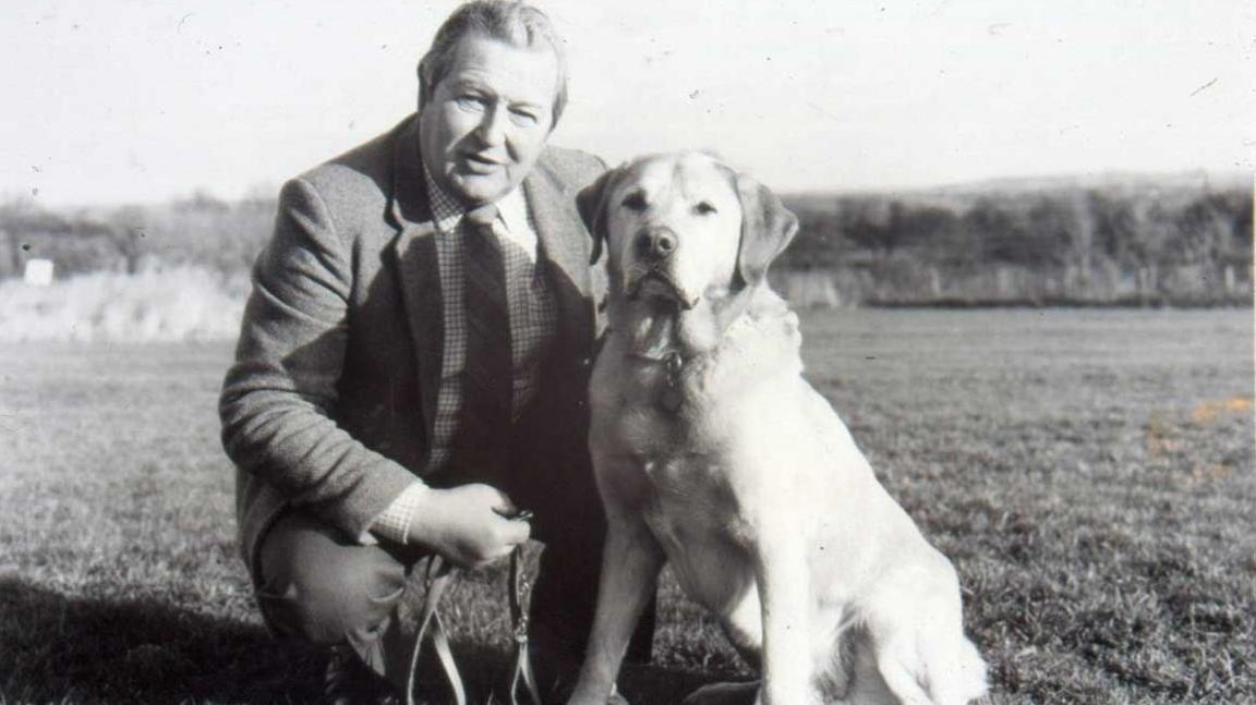 A black and white image of a man kneeling in a field with a Labrador dog. The man is wearing a suit, checked shirt and striped tie. The dog is on a lead.