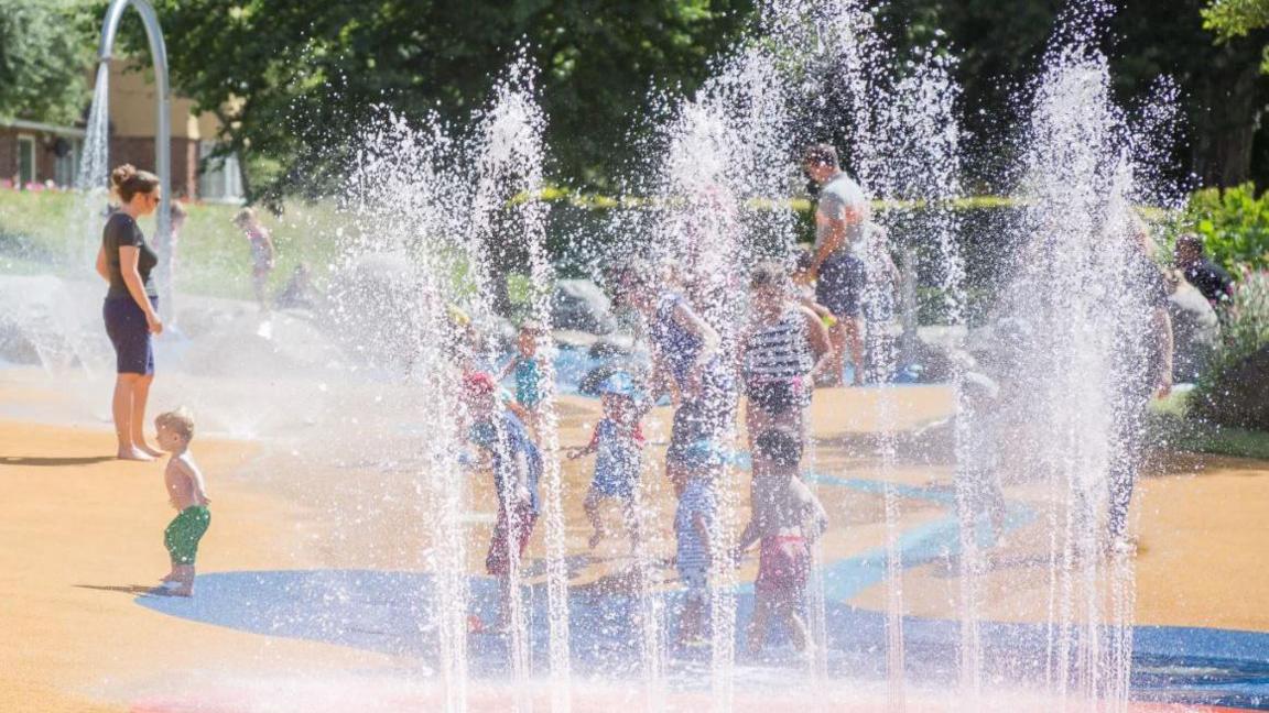 Children and parents play under water fountains at a park play area. There is a yellow and blue floor with water fountains dotted across it. There are lawns and trees in the background, as well as some houses that are partly visible on the edge of the park.