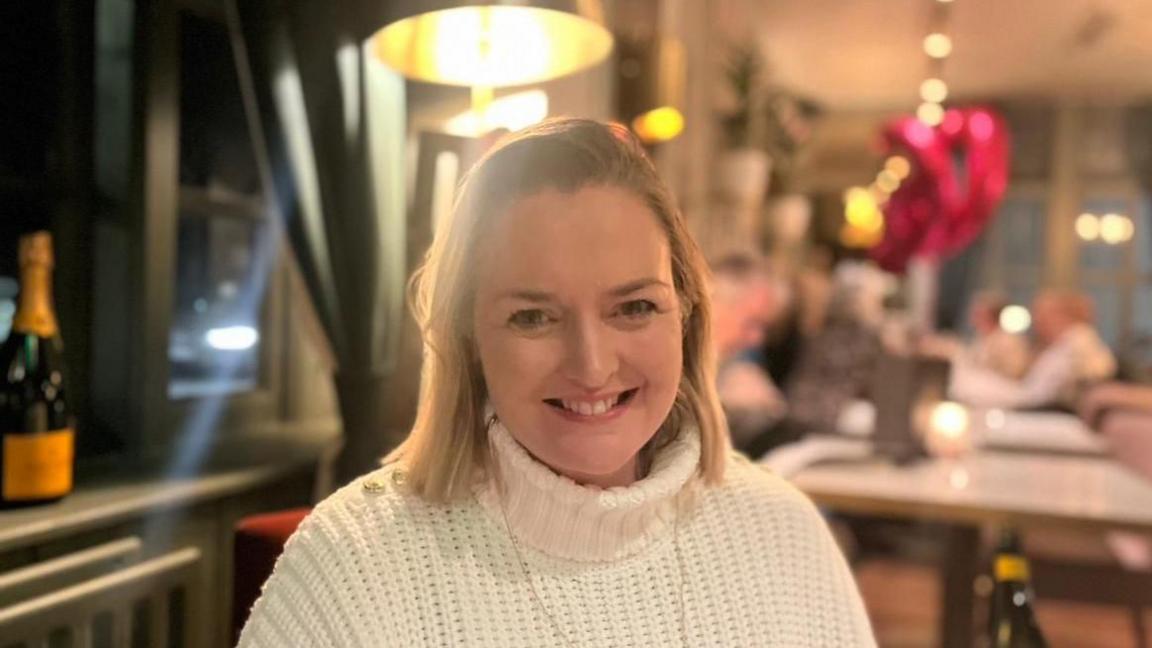 Picture of Natalie in a restaurant smiling and wearing a white roll neck sweater and small gold necklace.