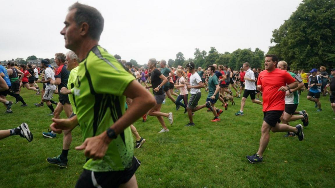 Dozens of people wearing shorts and t-shirts running from right to left across a park. In the foreground of the shot is a man wearing a green and black t-shirt and black shorts