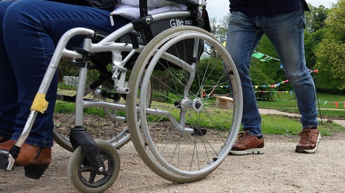 Bottom half of a person pushing another person in a wheelchair along a path in Ilam Park, in the village of Ilam, near Ashbourne, in Derbyshire.


