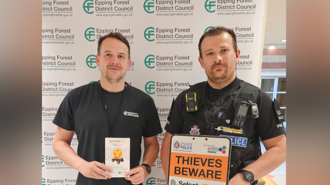 Epping Forest councillor Duncan Forsyth is pictured standing next to PC Warren Shepherd. Duncan has short dark hair and is wearing a black t-shirt with blue denim jeans. He is holding a property marking information leaflet. PC  Shepherd is wearing his officer uniform. He has dark short hair and is holding a sign warning thieves off.