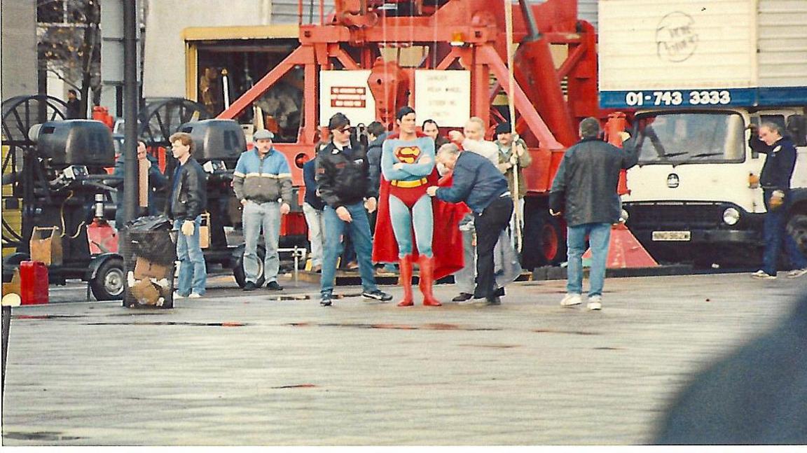 Christopher Reeve as Superman stands surrounded by crew members during filming in Milton Keynes. A red crane, industrial equipment and urban buildings are in the background.
