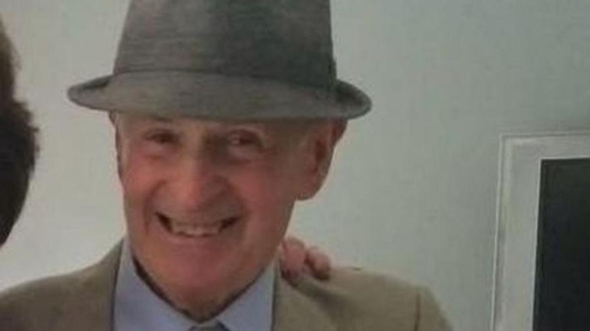 An elderly man smiles for a photo. He is wearing a grey hat and a cream jacket and light blue shirt.