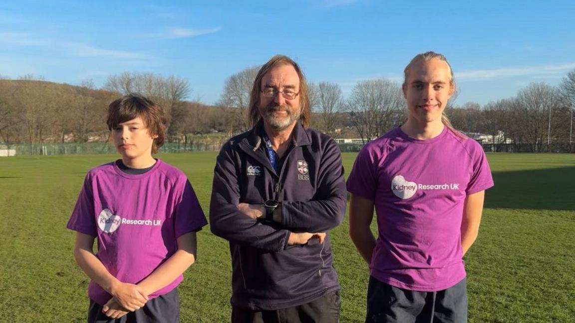 Two teenage boys wearing a purple t-shirt with a white logos and a man standing in the middle of them with long, brown hair and wearing a blue sports top. 