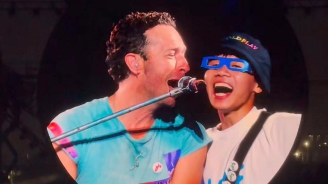 A circular screen at a concert shows Chris Martin who has short curly brown hair and a blue top singing into a mic and looking at a man to his side. The man next to him has his mouth wide open singing and is wearing rectangular blue glasses and a Coldplay bucket hat.