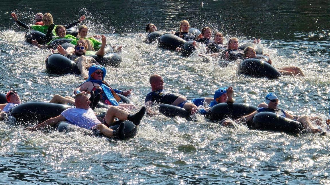 Teams of six in inflatable rings, which are linked together like a snake, racing in Castletown Harbour. The competitors are using their arms to paddle, which is creating lots of white splash. 