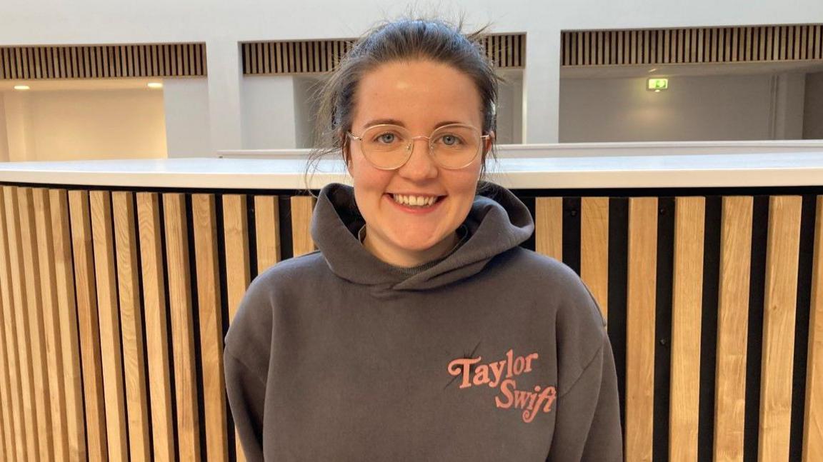 Ella Smith, with dark hair and glasses, is wearing a top with the words Taylor Swift on it. She is smiling at the camera.