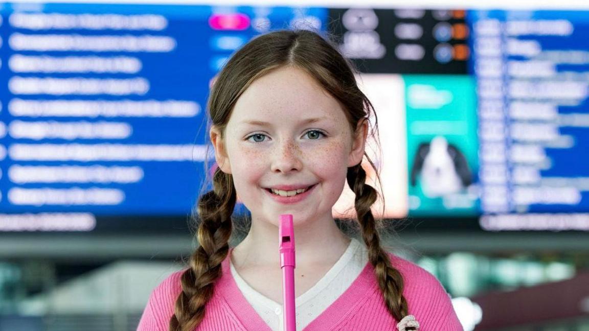 A young girl with freckles and two plaits is playing a pink tin whistle. She is wearing a pink top. The background is blurred but it looks like an arrivals and destination board at a train station.