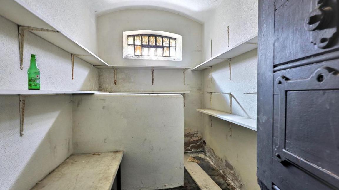 Inside view of one of the cells, there's a hard wooden bench on one side and a low wall beyond it obscuring the other side. Shelves line the wall, with a green glass bottle on one of them.