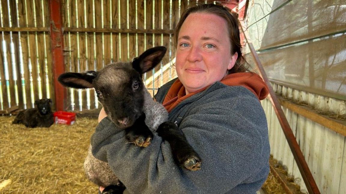 Steph Spiers holds an animal inside an area, with wooden planks as its perimeter. She is wearing a grey top and looking straight at the camera.