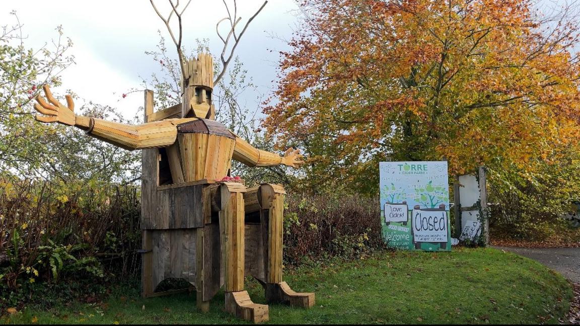 The Torm Man structure sitting with its arms outstretched. There is a pile of apples in its lap. It has been placed in front of a hedge near a sign that says "Torre Cider Farm".