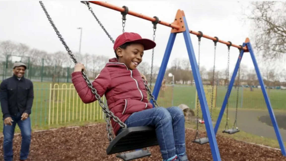 Boy smiling on the swing and his father behind smiling
