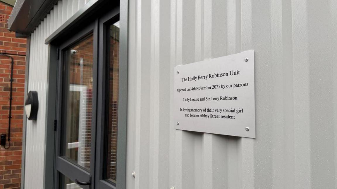 A metal cabin with a sign that reads: THE HOLLY BERRY ROBINSON UNIT OPENED ON 14TH NOVEMBER BY OUR PATRONS LADY LOUISE AND SIR TONY ROBINSON IN LOVING MEMORY OF THEIR VERY SPECIAL GIRL AND FORMER ABBEY STREET RESIDENT
