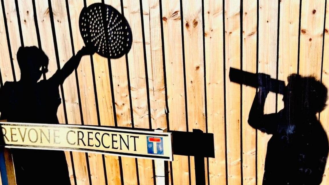 A wooden fence with two shadows cast upon it of people - one holding a cereal box to their eyes and peering through the opening, and the other holding a pizza tray with holes in above their head. There is a street sign at the bottom right of the image with the name 'Trevone Crescent' written on it.