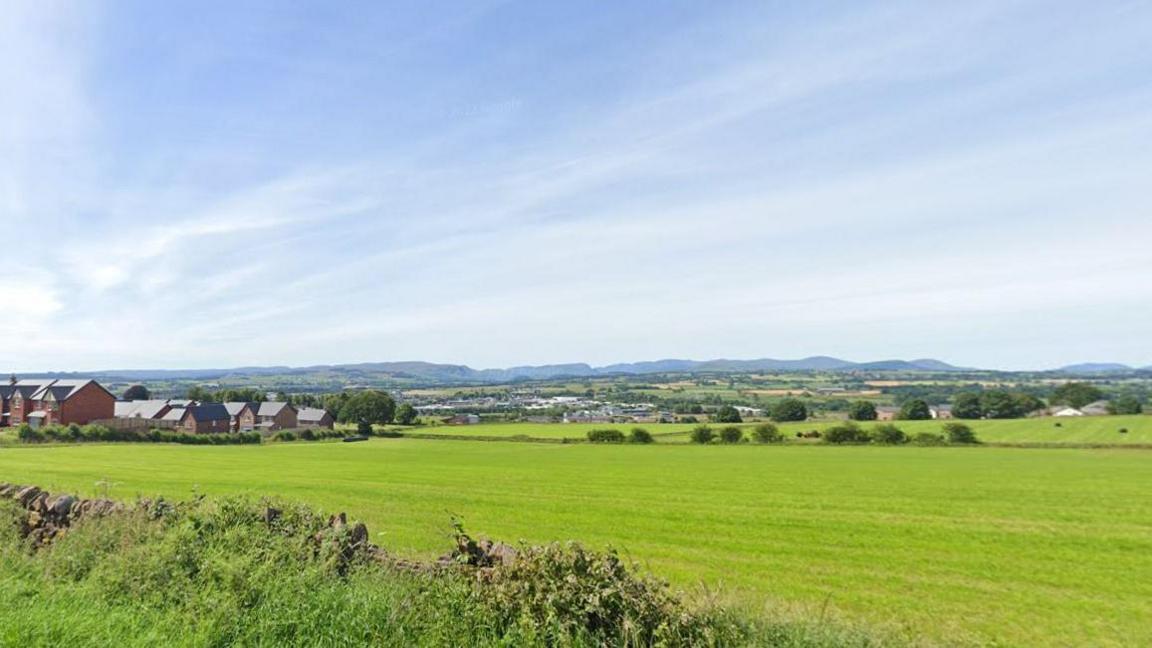 A blue sky and green field with houses on the edge of it. The area is surrounded by countryside