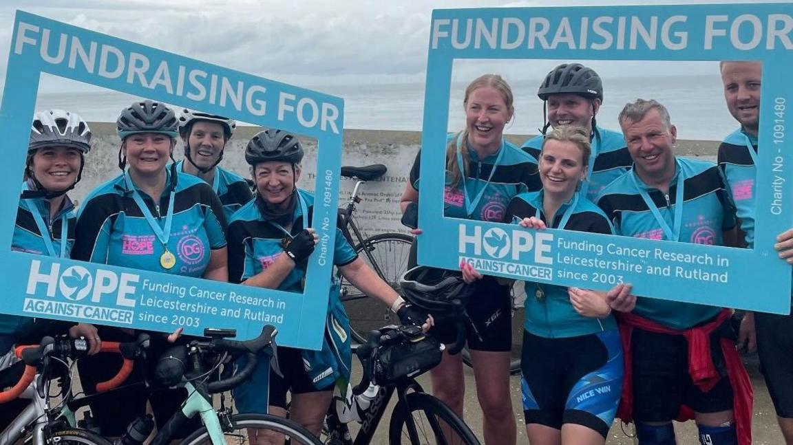 Two groups of people with bikes, dressed in cycling gear, look through square blue signs that say 'fundraising for Hope Against cancer'.