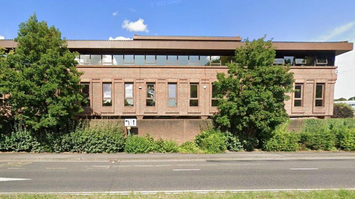 Two-storey light-brick office building with a row of small square windows below the roof and taller windows on the floor below. There are two trees in front of the building and a road in the foreground.