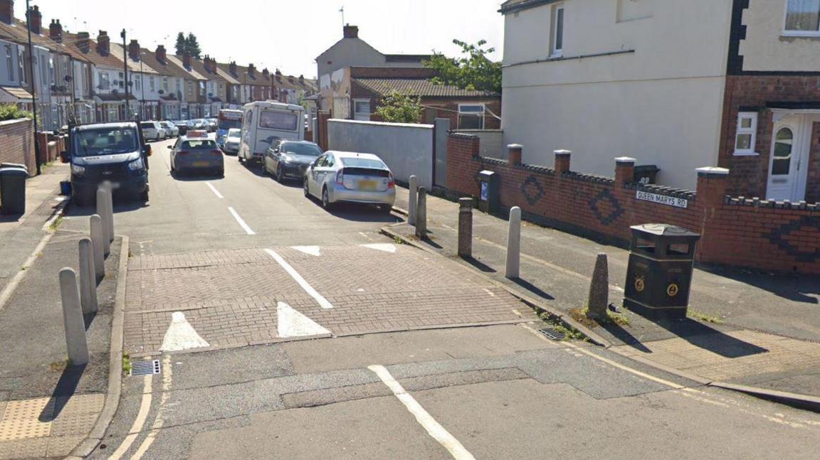 A Google street view image of Queen Mary's Road. The road sign is on a low red brick wall to the right and rows of terraced houses can be seen on both sides of the street, with cars parked outside them on the road.